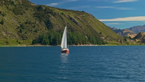 Low-aerial-orbit-around-sailboat-gliding-across-Lake-Hawea-reflecting-white-sail-in-water