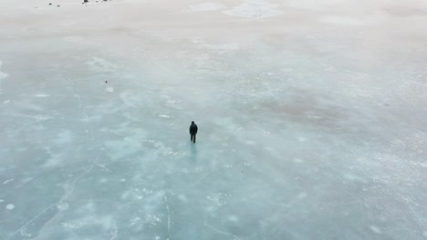 Toma-Aérea-De-Un-Hombre-Solitario-Caminando-Sobre-Hielo-Y-Bailando-En-Un-Vasto-Y-Vacío-Lago-Congelado-Cerca-De-Reikiavik,-En-Islandia