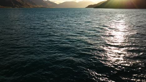 Sun-flare-rays-reflect-across-shimmering-waters-of-Lake-Wakatipu-with-silhouette-of-mountains