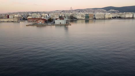 Leuchtturm-Bei-Sonnenuntergang,-Schwenk-Zur-Uferpromenade-Von-Thessaloniki:-Luftaufnahme-Einer-Drohne-Zeigt-Leuchtturm-Mit-Vögeln,-Im-Hintergrund-Die-Skyline-Der-Stadt-–-Faszinierende-Ausblicke-Auf-Die-Schönheit-Der-Griechischen-Stadtküste-Θεσσαλονίκη,-Ελλάδ?