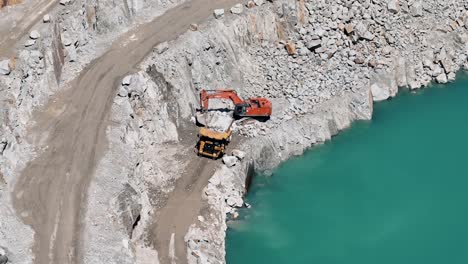 Excavadora-Llenando-Un-Camión-Volquete-En-Una-Cantera-Junto-A-Un-Sereno-Lago-Azul---Aéreo