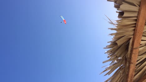 Straw-beach-umbrella,-blue-sky-and-peson-on-paraglide-with-Cuban-flag-wing