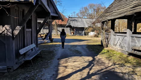 Woman-Walking-Through-Old-Wooden-Structures-With-Farmhouse-In-Traditional-Farming-Village