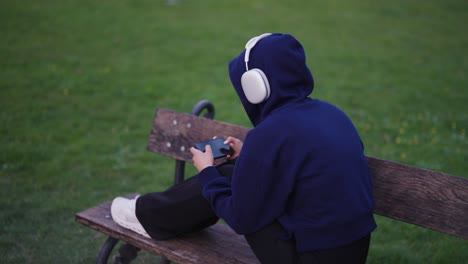 Lonely-woman-in-hoodie-with-headphones-sit-on-wooden-bench-and-use-smartphone