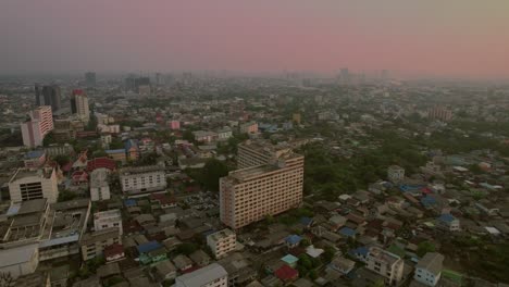 Aerial-drone-forward-moving-shot-over-the-city-buildings-in-Bangkok,-Thailand-with-orange-sky-after-sunset-in-the-background