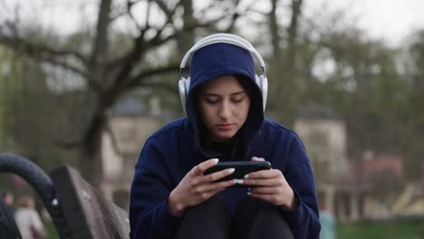 Sad-woman-in-hoodie-sit-on-outdoor-bench,-scroll-smartphone-with-headphones-on