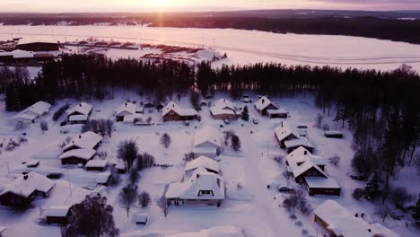 Aerial-winter-sunset-over-snow-covered-neighborhood-next-to-a-frozen-lake-and-a-forest