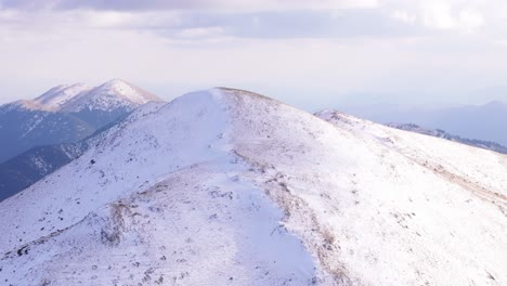 Snow-covered-mountain-peak-in-light-dusting-blankets-over-rocky-terrain,-aerial-descending