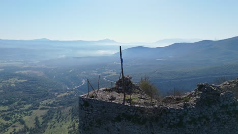 Flagpole-with-rolled-flag-blowing-lightly-in-wind-at-edge-of-Karytaina-Castle-in-Peloponnese-Greece,-aerial-pullback-to-village