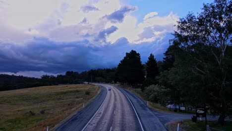 Reverse-drone-shot-of-white-car-driving-along-rural-highway-with-dark-storm-clouds-on-the-horizon-at-Crackenback,-NSW,-Australia