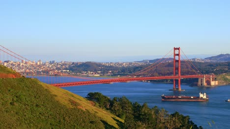 Die-Golden-Gate-Bridge-Von-Einem-Malerischen-Aussichtspunkt-Mit-Blick-Auf-Die-Bucht,-Unter-Der-Ein-Frachtschiff-Bei-Sonnenuntergang-Mit-Warmem-Licht-Und-Blauem-Himmel-Hindurchfährt