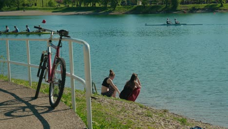 Dos-Amigos-Sentados-Junto-A-Las-Serenas-Aguas-Del-Lago-Jarun,-Zagreb,-Con-Una-Bicicleta-Estacionada-Cerca