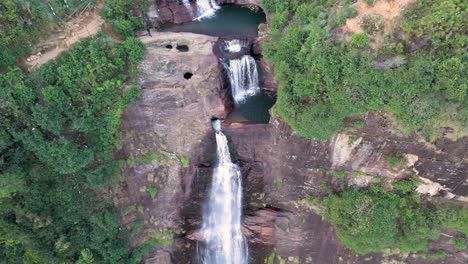 Breathtaking-drone-shot-of-Gartmore-Falls-in-Sri-Lanka,-featuring-lush-greenery,-orange-toned-sands,-and-rocks-against-a-vivid-blue-sky