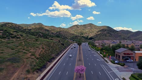 Drone-Flight-over-Incoming-Traffic-along-Railroad-Canyon-Road-in-Lake-Elsinore-California-with-Palm-Trees-in-the-Center-Median-Clouds-in-sky-and-casting-shadows