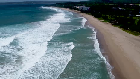 Waves-lapping-on-a-sandy-beach-with-lush-trees,-daylight,-aerial-view