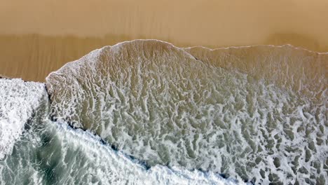 Schaumige-Wellen-Brechen-An-Einem-Sandstrand,-Die-Raue-Schönheit-Der-Natur-Von-Oben-Eingefangen,-Luftaufnahme