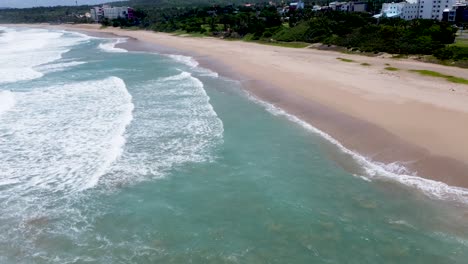 Aerial-shot-of-waves-gently-breaking-on-the-shore-of-a-secluded-beach,-lush-greenery-in-the-background