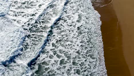 Frothy-ocean-waves-washing-onto-a-sandy-shore,-aerial-view