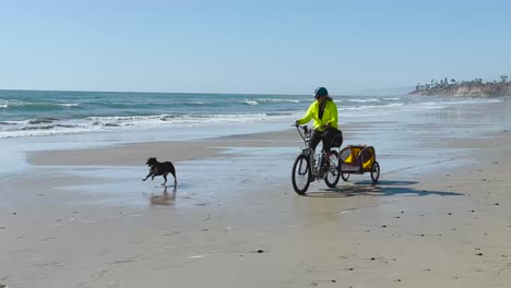 White-female-riding-bike-on-the-beach-with-a-dog-during-a-beautiful-sunny-day