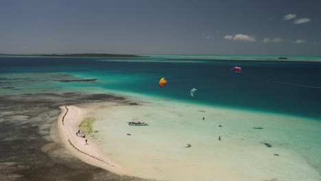 Kitesurfistas-Deslizándose-Sobre-Aguas-Cristalinas-De-Color-Turquesa-Por-Un-Islote-Arenoso,-Los-Roques,-Vista-Aérea