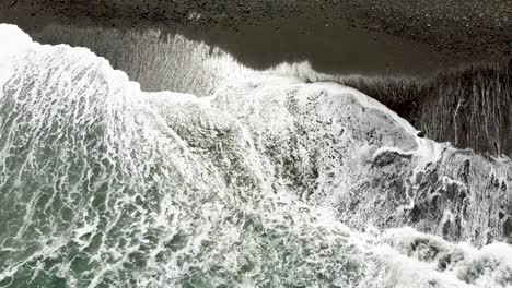 Waves-breaking-on-a-dark-sandy-beach,-contrasting-foam-and-textures,-aerial-view