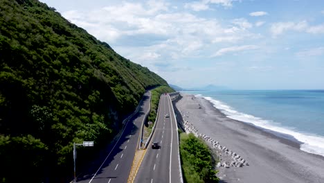 Coastal-road-curving-along-a-lush-hillside-and-beach,-blue-sea-on-a-sunny-day,-aerial-shot