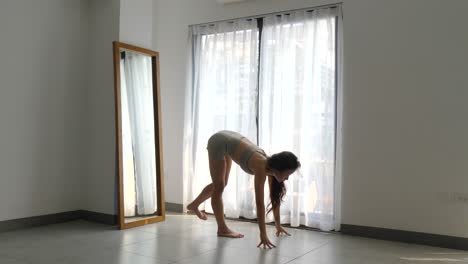 Mujer-Practicando-Yoga-Matutino-En-Una-Habitación-Iluminada-Por-El-Sol,-Estirándose-Con-Una-Expresión-Serena,-Espejo-A-Un-Lado