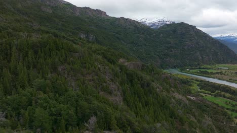 Aerial-Flying-Over-Forested-Hillside-To-Beside-Futaleufu-River-With-Snow-Capped-Mountains-In-Background