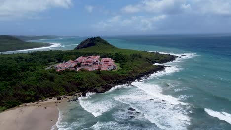 Coastal-resort-on-a-cape-with-crashing-waves,-sunny-with-some-clouds,-aerial-view