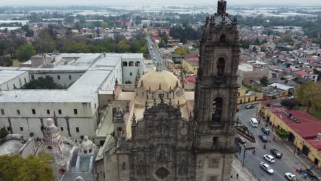 Eine-Drohnenaufnahme-Einer-Basilika-In-Tepoztlan-Mit-Einigen-Straßen-Und-Autos,-Die-An-Einem-Bewölkten,-Sonnigen-Tag-Vorbeifahren