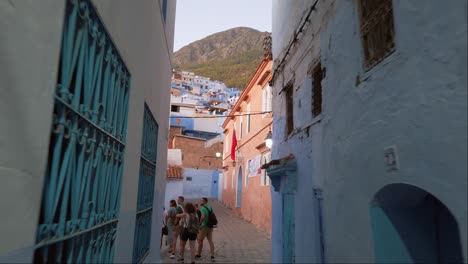 A-group-of-tourists-walks-through-a-narrow-alley-in-the-town-centre-of-Chefchaouen,-with-blue-colored-terraced-houses-and-the-Rif-Mountains-emerging-in-the-background-in-Morocco