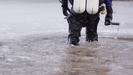 Geared-up-ice-diver-in-dry-suit-walks-into-water-and-sits-on-edge-of-lake-ice