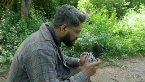 Professional-drone-pilot-using-DJI-drone-remote-controller-with-a-smartphone-attached-in-a-forest-landscape-for-wildlife-surveillance,-India