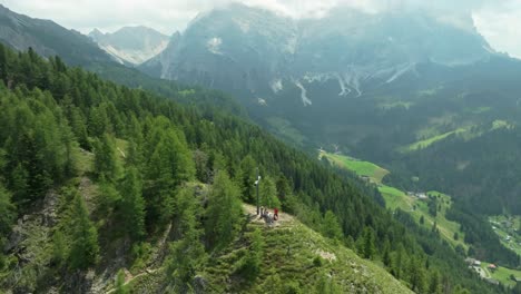 Swift-aerial-orbit-view-captures-the-cross-atop-Crucs-da-Rit-peak,-with-La-Val-village-and-the-majestic-Dolomites-in-the-backdrop