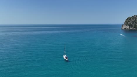 Velero-Navegando-Por-Las-Aguas-Turquesas-Cerca-De-La-Costa-Rocosa-De-La-Isla-De-Corfú,-Grecia,-Bajo-Un-Cielo-Azul-Claro