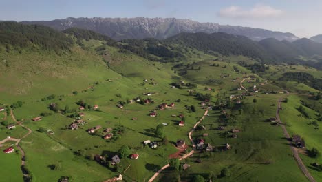 A-scenic-village-nestled-in-green-hills-with-mountain-backdrop,-under-clear-skies,-aerial-view