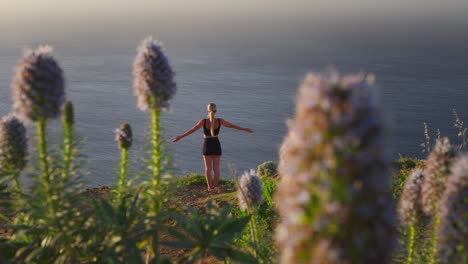 Woman-raising-arms-meditating-on-cliff-overlooking-sea,-Pride-of-Madeira-flower-bokeh