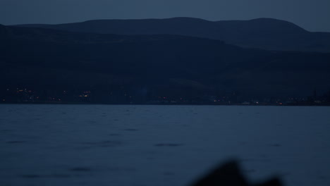 Coastal-town-lights-twinkle-in-distance-at-base-of-mountains-across-water