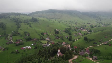 A-foggy-village-with-a-church-amidst-green-hills,-aerial-view