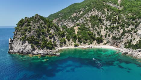Speedboat-cruising-near-the-lush-cliffs-of-Corfu-Island-on-the-turquoise-Ionian-Sea,-sunny-day,-aerial-view