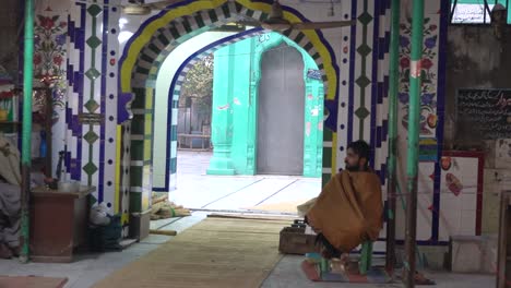 Man-wrapped-in-a-blanket-sitting-at-the-colorful-entrance-of-a-shrine-in-Gujrat,-Pakistan