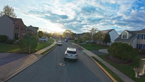 Drone-Siguiendo-La-Toma-De-Un-Auto-Plateado-En-La-Carretera-En-Una-Zona-Residencial-Americana-Durante-La-Puesta-De-Sol