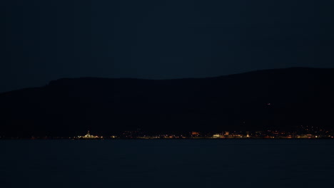 Coastal-town-lights-up-at-night-with-ferry-boats-crossing-paths-in-front-below-mountains