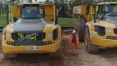 Mit-Persönlicher-Schutzausrüstung-Ausgestatteter-Bauarbeiter-Inspiziert-Eine-Flotte-Von-Muldenkippern-Auf-Einer-Baustelle