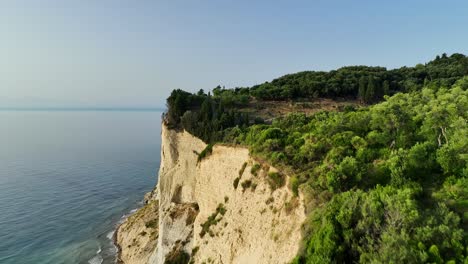 Sun-drenched-cliffs-of-Corfu-Island-overlooking-the-Ionian-Sea,-lush-greenery-encasing-the-landscape,-aerial-view