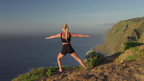 Woman-leaning-into-warrior-yoga-pose-on-cliff-at-sea-during-golden-hour