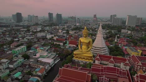 Una-Gigantesca-Estatua-Dorada-De-Buda-En-El-Bangkok-Urbano-Al-Atardecer,-Atmósfera-Serena,-Vista-Aérea
