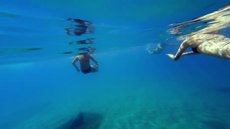 Two-women-in-black-bikinis-are-swimming-in-a-clear-blue-ocean-with-the-sun-shining-down,-with-the-camera-submerged-in-the-water-just-below-the-surface,-and-facing-two-women-swimming-away-from-it
