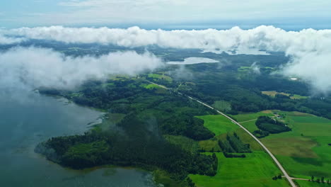 Lush-green-landscape-with-lake,-roads,-and-low-clouds,-daylight,-aerial-view