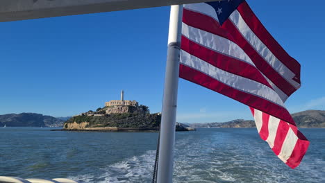 Bandera-Americana-Ondeando-En-Ferry-Con-La-Isla-De-Alcatraz-Y-Edificios-Penitenciarios-En-El-Fondo,-San-Francisco-CA,-EE.UU.